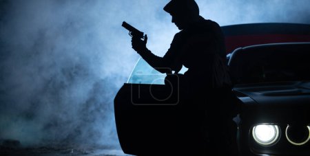 Photo for Silhouette of the Bandit with a Gun Next to His Muscle Car in the Night Fog. Dark Scenery. Outlaw Life Theme. - Royalty Free Image
