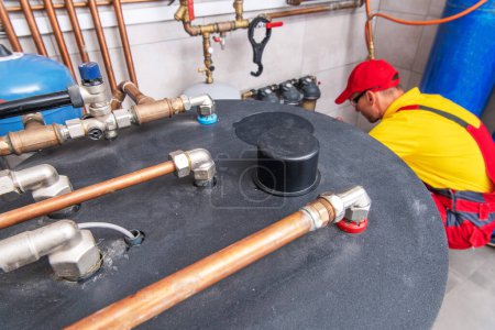 Foto de Aerial View of Tank-Type Hot Water Heater Inside Residential Boiler Room. Plumber Performing Heating System Maintenance in the Background. - Imagen libre de derechos