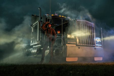Foto de American Semi Truck Driver and His Vehicle During Foggy Night. Ground Shipping Transportation. Automotive Industry. - Imagen libre de derechos