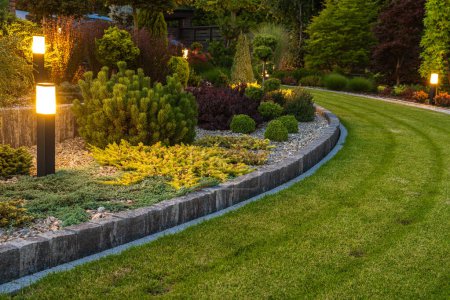 Foto de Professionally Landscaped Backyard Garden with Evenly Mowed Lawn and Trimmed Shrubs Illuminated with Outdoor Bollard Lamps. - Imagen libre de derechos