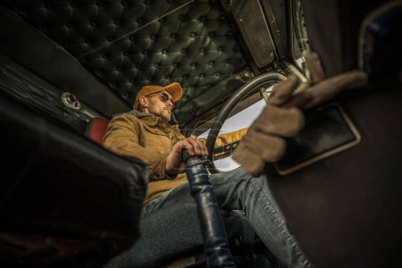 Téléchargez les photos : Camionneur caucasien professionnel conduisant son semi-camion américain. Vue du bas de l'intérieur de la cabine. - en image libre de droit
