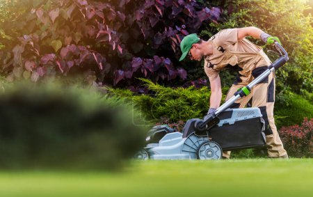 Foto de Caucasian Garden and Landscaping Worker Mowing  Backyard Lawn Using Electric Cordless Grass Mower. Garden Maintenance Theme. - Imagen libre de derechos