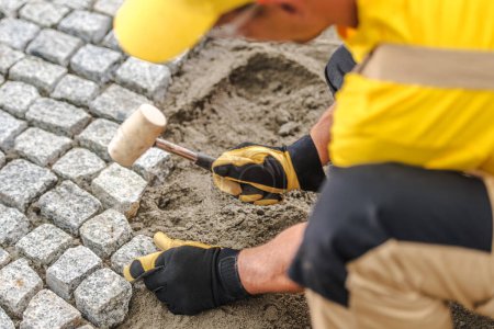 Photo for Closeup of Professional Contractor in Yellow Uniform Laying Paving Stones Pounding Them Into the Sand. Home Improvement Theme. - Royalty Free Image