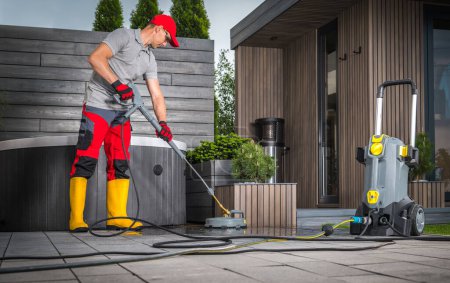 Caucasian Man in His 40s Pressure Washing Concrete Bricks Patio Using Surface Attachment. Modern Garden Shed and a Hot Tub in a Background.