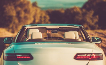 Téléchargez les photos : Vue arrière de la voiture convertible blanche moderne avec toit rétractable dans le coucher des lumières du soleil. Blanc mâle assis dans le siège du conducteur. - en image libre de droit