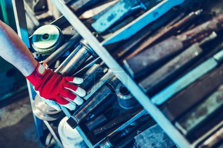 Foto de Mano de trabajador industrial en guante rojo tocando herramientas y brocas industriales de servicio pesado en el estante del taller. - Imagen libre de derechos