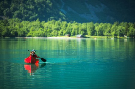 Foto de Hombre caucásico en canoa roja cruzando el lago glacial en Noruega. Disfrutando del soleado Día de Noruega. - Imagen libre de derechos
