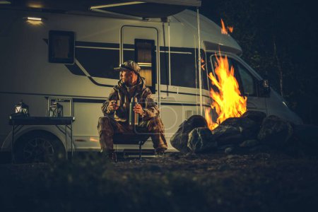 Foto de Hunter con recubrimiento de camuflaje Burning Campfire junto a su autocaravana Camper Van Recreational Vehicle. Tema de temporada de caza. - Imagen libre de derechos