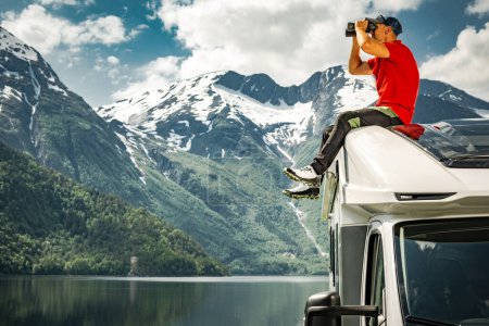 Hombre caucásico de 40 años explorando la escénica naturaleza noruega usando prismáticos mientras está sentado en el techo de su vehículo recreativo Camper Van RV. Noruega, Europa.