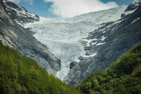 Foto de Scenic Mountainscape of Vestland County in Norway (en inglés). Montaña rocosa cubierta de nieve y rodeada de un denso bosque verde. - Imagen libre de derechos