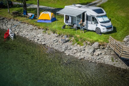 Luftaufnahme eines Reisemobilreisenden, der vor seinem Wohnmobil Kaffee genießt, nachdem er einen Campingplatz am Fluss eingerichtet hat. Thema Camping und Reisen.