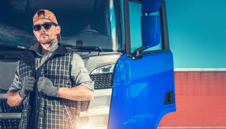 Foto de Conductor de camión caucásico delante de un camión semirremolque azul. Usar gafas de sol y guantes de protección. - Imagen libre de derechos