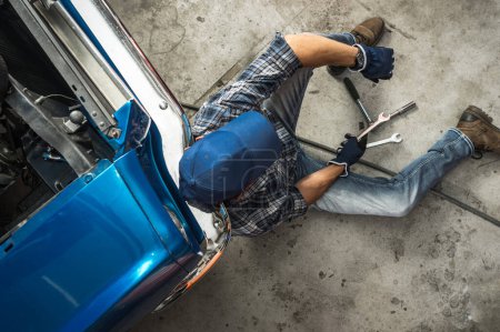 Photo for Tired Classic Cars Mechanic Resting on a Garage Floor Next to His Broken Muscle Car. Automotive Theme. - Royalty Free Image
