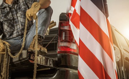 Téléchargez les photos : Rancher américain avec une corde assise sur son lit de camionnette à côté du drapeau national américain - en image libre de droit