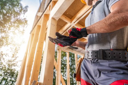 Téléchargez les photos : Entrepreneur de construction portant des gants de protection tout en restant à côté du cadre squelette de la maison en bois - en image libre de droit