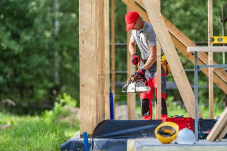 Téléchargez les photos : Travailleur du chantier de construction caucasien avec une tronçonneuse à l'intérieur cadre squelette en bois - en image libre de droit