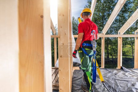 Téléchargez les photos : Entrepreneur de construction professionnel portant un harnais de sécurité à l'intérieur d'un cadre squelette en bois nouvellement construit - en image libre de droit
