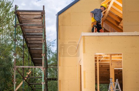 Téléchargez les photos : Travailleur de la construction et la petite maison résidentielle en bois nouvellement construite. Échafaudages à côté de la maison. - en image libre de droit