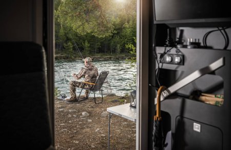 Téléchargez les photos : Homme caucasien détendu pêche à la mouche devant son camping-car Motor Home. Camping d'été. - en image libre de droit