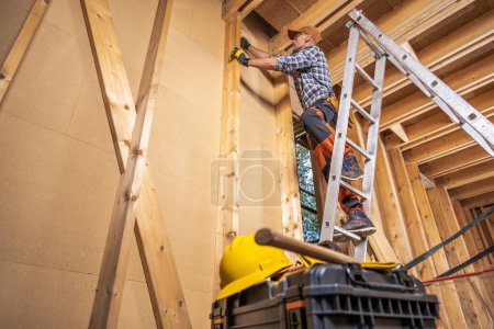 Téléchargez les photos : Entrepreneur de construction sur une échelle à l'intérieur d'une maison de structure en bois nouvellement développée - en image libre de droit