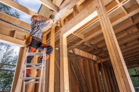 Téléchargez les photos : Bâtiment à ossature de bois de maison par le travailleur entrepreneur. Utilisation du marteau tout en restant sur une échelle - en image libre de droit