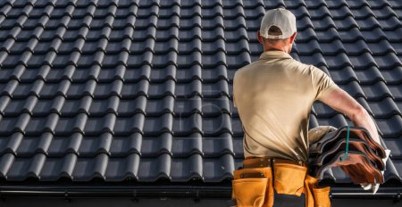 Foto de Trabajador profesional del contratista de techos caucásico moviendo un tejado negro baldosas de cerámica en sus manos - Imagen libre de derechos