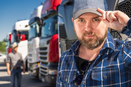 Foto de Camionero caucásico en sus 30 años en un sombrero de pie delante de un grupo de camiones de estilo euro. - Imagen libre de derechos