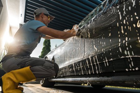 Foto de Un hombre está usando un spray de agua para lavar un auto. El hombre se centra en la limpieza del vehículo a fondo, rociando agua en el exterior de los coches para eliminar la suciedad y la suciedad. - Imagen libre de derechos