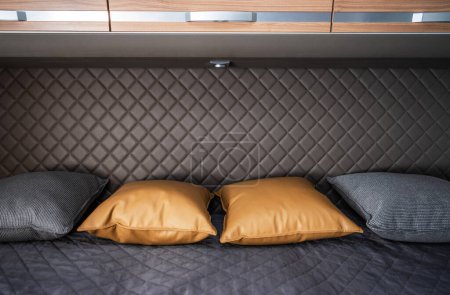 A close-up shot of a bed in a modern RV camper van. The bed is made with dark gray linens and features three pillows. Two pillows are made of a brown faux leather material and a single pillow is made of gray material. Above the bed is a wooden cabine