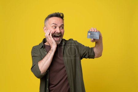 Photo for Astonished excited mature man with hand gesture at head in casual green shirt isolated on yellow background looking and showing bank plastic credit card holding in hand. - Royalty Free Image