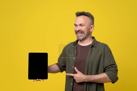 Happy smiled bristle and mustached mature man dressed in green shirt and brown t-shirt pointing with thumb up and finger to tablet pc gadget with black screen holding in hand on yellow background