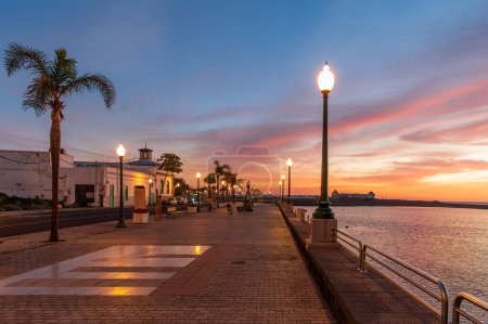 Téléchargez les photos : Promenez-vous sur la promenade sereine d'Arrecife au crépuscule à Lanzarote, où les silhouettes de palmiers et un coucher de soleil vibrant créent la toile de fond photo de stock parfaite. - en image libre de droit