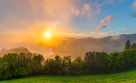 Genießen Sie den ruhigen Sonnenuntergang vom Miradouro do Pico dos Bodes, wo die Sonnenstrahlen die grünen Landschaften Sao Miguels in sanftem Nebel erleuchten.