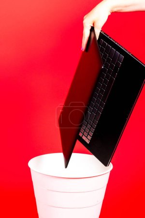 Téléchargez les photos : Ordinateur portable noir dans une poubelle sur fond rouge isolé - en image libre de droit