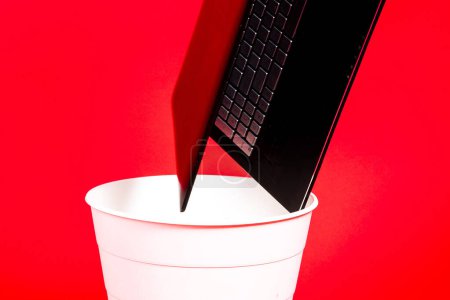 Black laptop in a trash bin on red background isolated