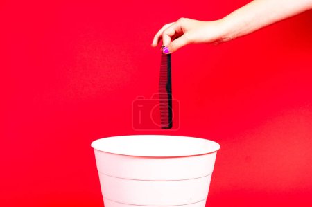 Hand throws plastic comb into trash bin with package on a red background