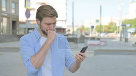 Foto de Molesto hombre de mediana edad reaccionando a la pérdida en el teléfono inteligente al aire libre - Imagen libre de derechos