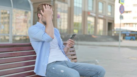 Foto de Hombre de mediana edad reaccionando a la pérdida en el teléfono inteligente mientras está sentado al aire libre en el banco - Imagen libre de derechos