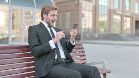 Foto de Businessman Reacting to Online Failure via Smartphone while Sitting Outdoor on Bench - Imagen libre de derechos