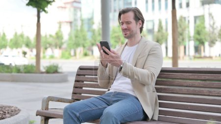 Foto de Man Reacting to Loss on Smartphone while Sitting on Bench - Imagen libre de derechos