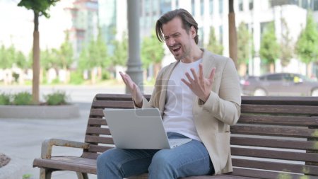 Foto de Man Reacting to Loss on Laptop while Sitting on Bench - Imagen libre de derechos