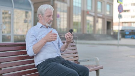Foto de Senior Old Man Reacting to Loss on Smartphone while Sitting Outdoor on Bench - Imagen libre de derechos