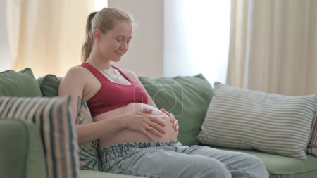 Photo for Pregnant Woman Stokes Her Big Belly while Sitting on Sofa - Royalty Free Image