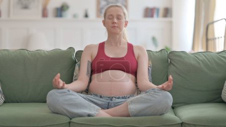 Téléchargez les photos : Meditating Pregnant Woman Doing Yoga while Sitting on Sofa - en image libre de droit