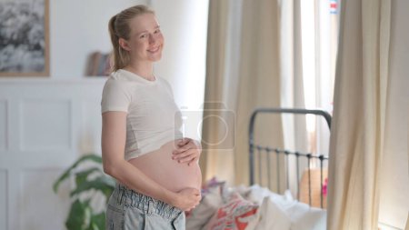 Téléchargez les photos : Standing Pregnant Woman Smiling at Camera while Holding Her Tummy - en image libre de droit