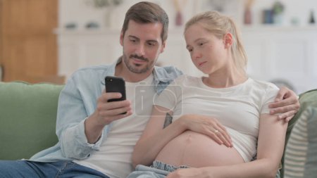 Foto de Pregnant Couple Using Smartphone while Sitting on Sofa - Imagen libre de derechos
