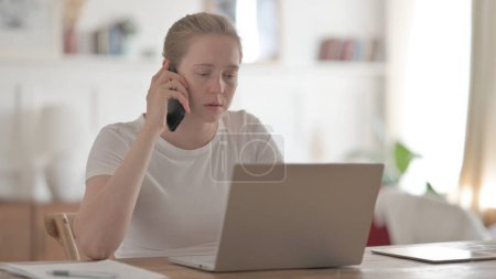 Téléchargez les photos : Jeune femme parlant au téléphone tout en utilisant un ordinateur portable au bureau - en image libre de droit
