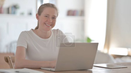 Téléchargez les photos : Jeune femme souriant à la caméra tout en utilisant un ordinateur portable au bureau - en image libre de droit