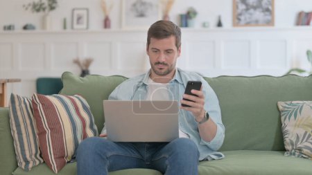 Foto de Hombre casual con ordenador portátil usando Smartphone en el sofá - Imagen libre de derechos