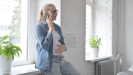 Téléchargez les photos : Enceinte belle femme parlant au téléphone tout en étant assis dans la fenêtre - en image libre de droit